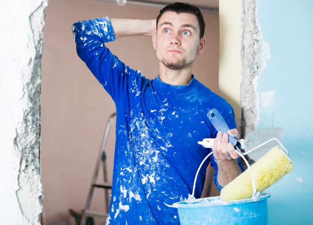 Man in a blue paint-splattered shirt looking puzzled while holding painting tools, illustrating the challenge of how to get paint out of your hair.