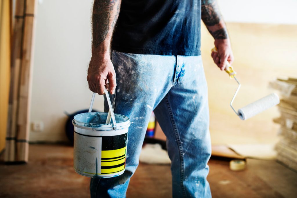 Painter wearing paint-stained jeans while holding a paint can and roller – how to get paint out of jeans including dried, latex, and acrylic paint.