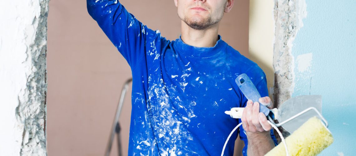 Man in a blue paint-splattered shirt looking puzzled while holding painting tools, illustrating the challenge of how to get paint out of your hair.